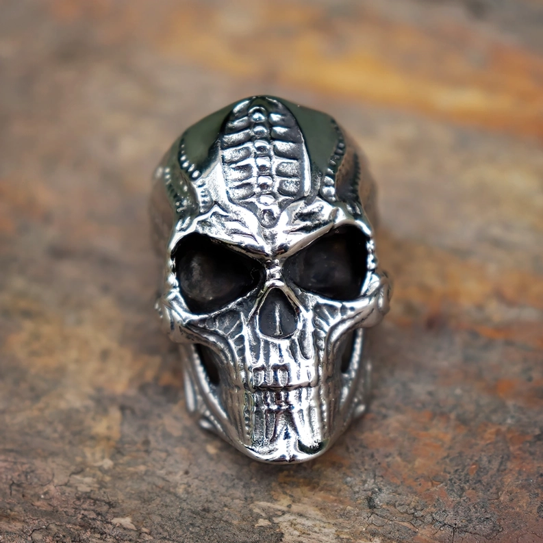 Biomechanischer gotischer Totenkopf-Ring in Frontansicht auf rustikalem Stein mit eingravierter Totenkopf-Details und silbriger Metallfinish