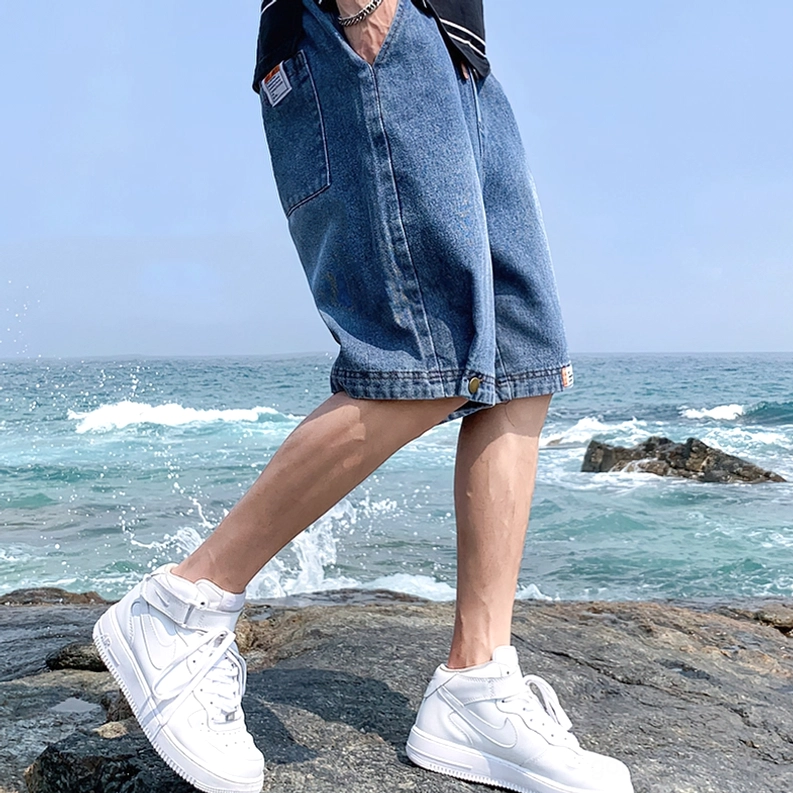 Männliches Modell in blauen Gothic lockeren Denim Shorts Drei-Viertel-Seitenwinkel an Küstenlandschaft – Blaue lockere Shorts Seitenansicht