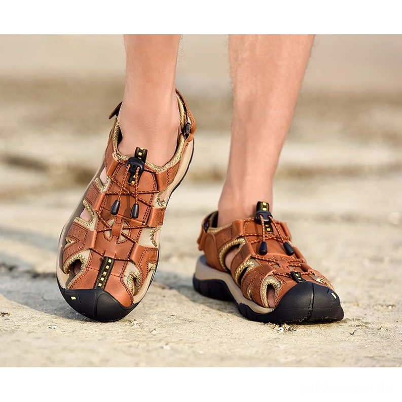 Braune gotische Knöchelriemen-Sandalen getragen auf männlichem Bein auf gepflastertem Boden, praktische dunkle Alternative Alltagsstil-Szene