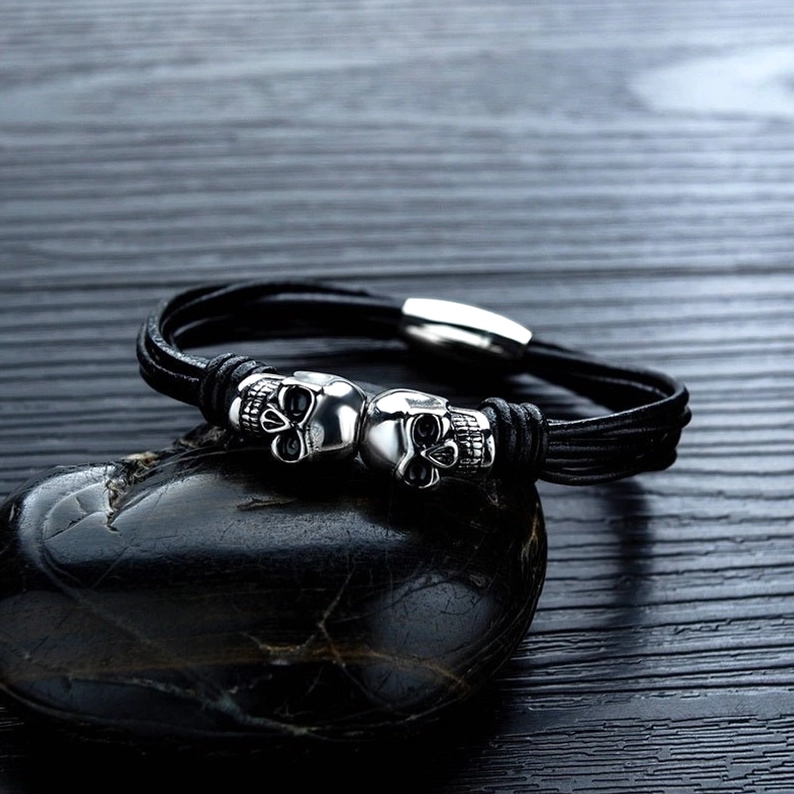 Seitenansicht des Doppelschädel Gothic Armbandes auf dunkler Holzoberfläche mit schwarzem Stein-Styling-Zubehör, zeitlose gotische Ästhetik