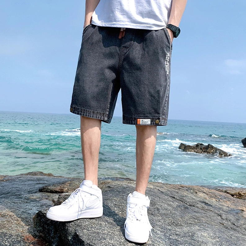Männliches Modell trägt dunkelgraue Gothic Denim Shorts mit weißen Sneakern in Küstenumgebung, Vorderansicht – Herren Gothic Shorts Strandstyling