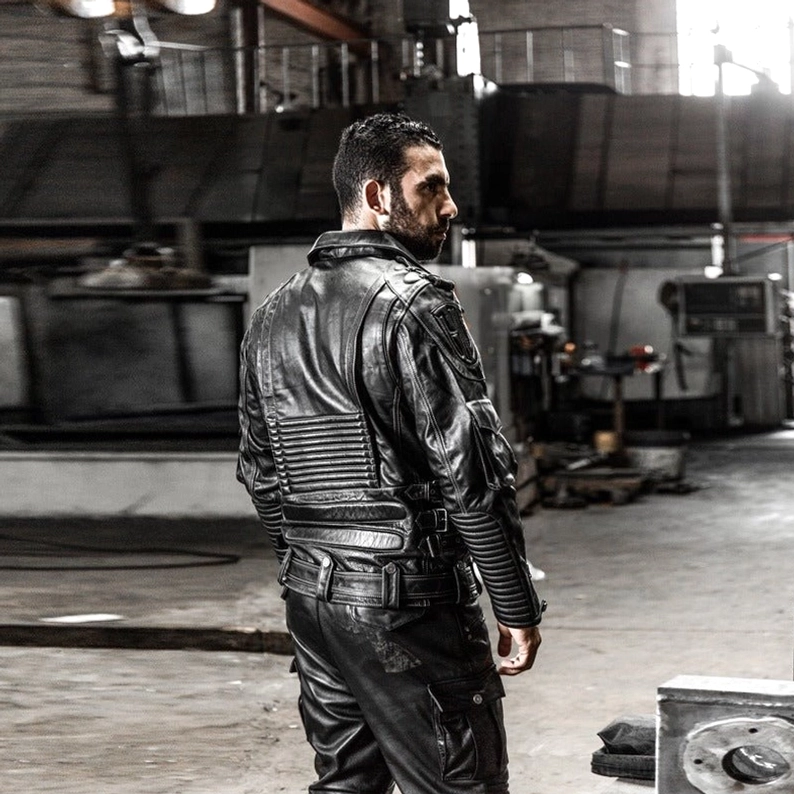 Herrenmodell trägt schwarze echte Leder Gothic Motorradjacke von hinten, gerippte Rückenpaneele sichtbar, Taillenriemen-Schnallen deutlich, Rückensilhouette, Industrial-Hintergrund