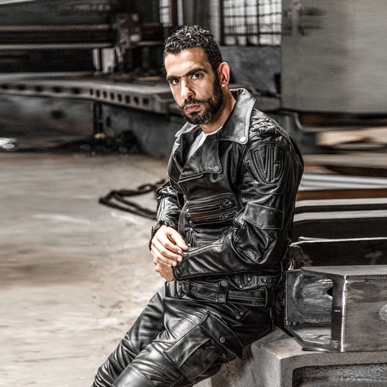 Herrenmodell trägt schwarze echte Leder Gothic Motorradjacke und Lederhose, entspannte sitzende Pose, industrielle Ästhetik, kompletter schwarzer Biker-Outfit, Seitenprofil