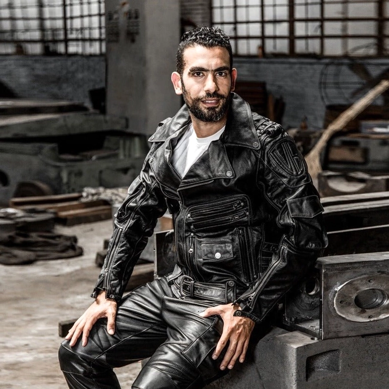 Herrenmodell trägt schwarze echte Leder Gothic Motorradjacke, eng anliegende Rider-Silhouette, industrielle Werkstatt-Kulisse, vollständiges Gothic-Outfit-Styling, Frontalaufnahme