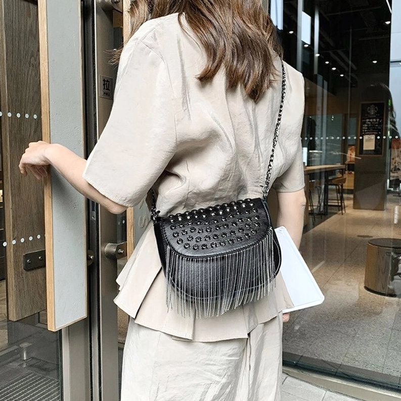 Frau trägt schwarze gotische Nieten-Schultertasche mit Fransen im Crossbody-Stil, Rückansicht in indoor Café-Umgebung zeigt die elegante Gothic-Umhängetasche
