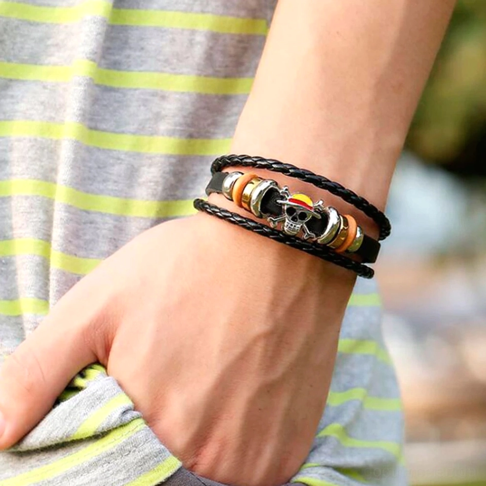 Schwarzes geflochtenes Leder Gothic Schädel Armband am männlichen Handgelenk getragen – mehrschichtige Lederwickel-Design zeigt Punk-Ästhetik in alltäglicher Outdoor-Umgebung mit graugestreiftem Oberteil