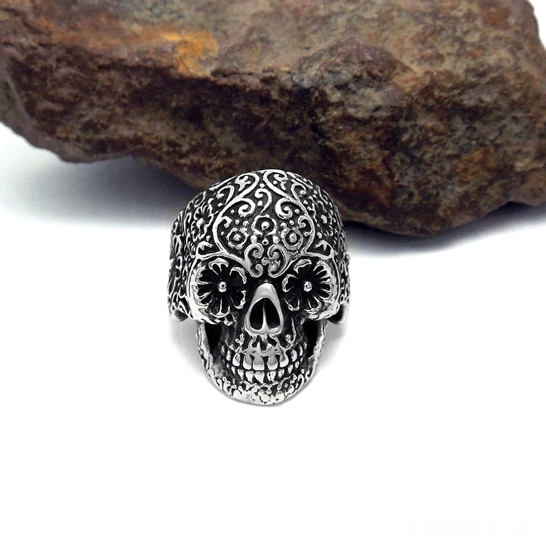 Dunkler silberner gotischer Totenkopfring auf rustikalem Steinuntergrund, detaillierte geschnitzte Blumenmuster auf dem gesamten Schädel, Punk-Rock-Alternative Schmuckstücke mit antiker Patina