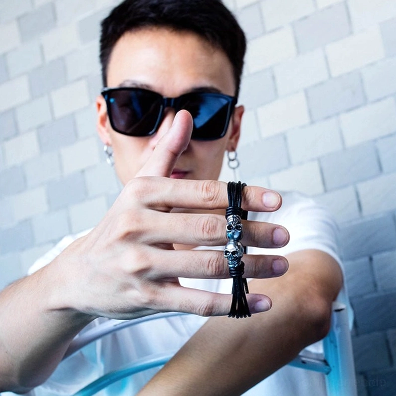 Mann hält Doppelschädel Gothic Armband in der Hand mit Quaste-Detail, Herren Schmuck Nahaufnahme auf gefliesten Innen-Hintergrund