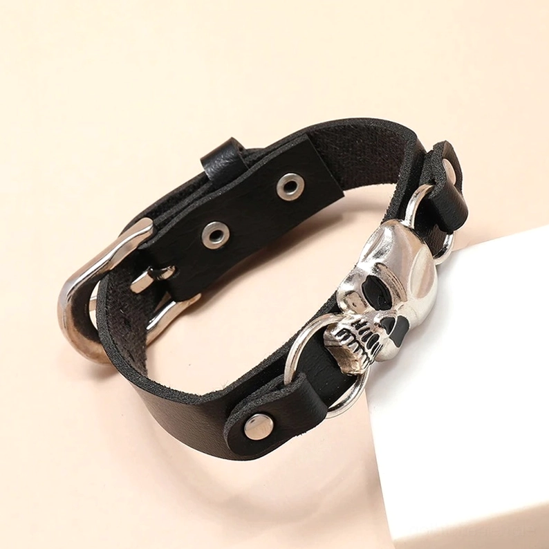 Schwarzes Leder Totenkopf-Armband mit silberner Metallschließe, Tiefenwinkel 3/4 Ansicht auf pfirsichfarbenem Hintergrund, Gothic Manschettenarmband
