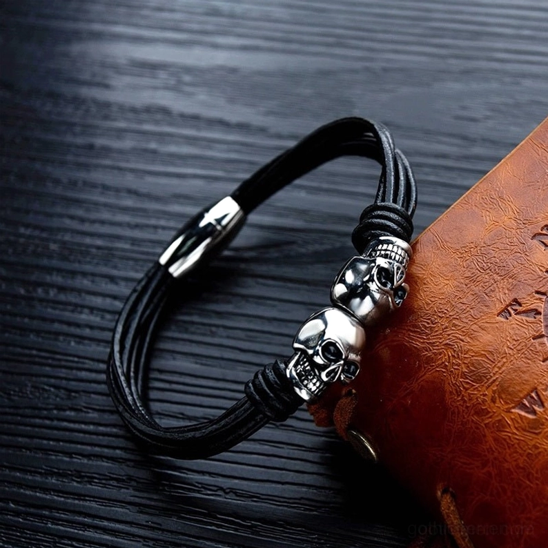 45-Grad-Winkelansicht des Doppelschädel Edelstahl Gothic Armbandes mit schwarzem Leder-Notizbuch-Styling auf Holz-Hintergrund, Memento-Mori