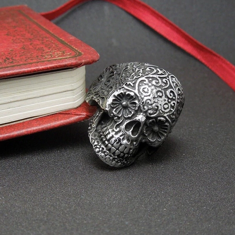 Seiten-Vorderansicht des Memento-Mori-Totenkopfrings mit floraler Dekoration auf dunkler Oberfläche, gotische Schmuckästhetik mit detaillierter Handengraving, professionelle Produktfotografie