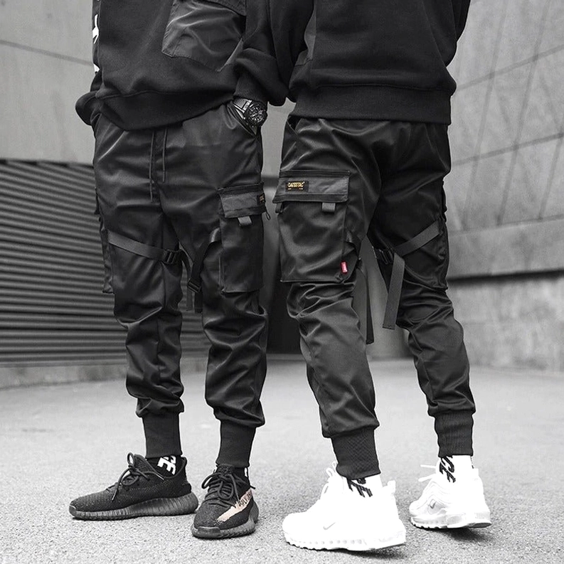 Zwei männliche Models in schwarzen Cargo Joggern in industrieller Outdoor-Umgebung, zeigt vielseitige Gothic-Streetwear-Styling-Möglichkeiten mit verschiedenen Sneaker-Styles