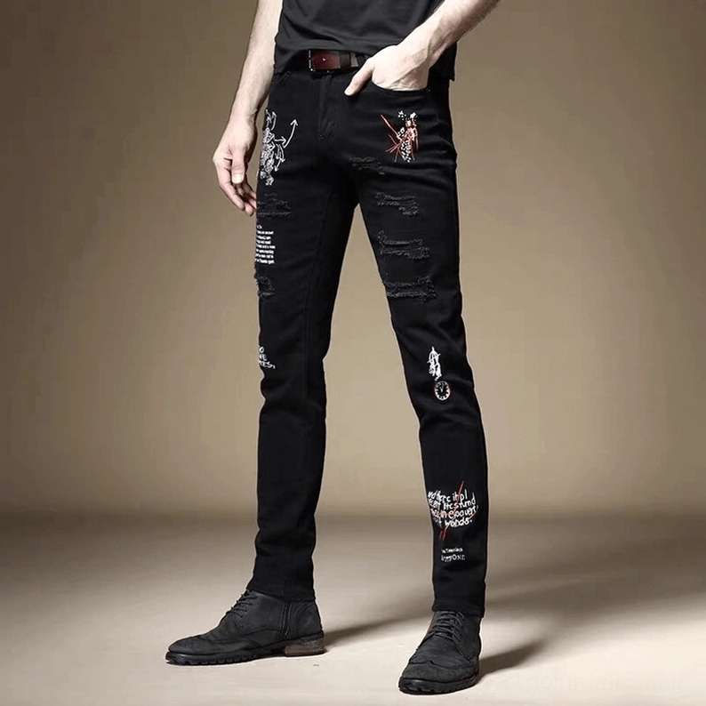 Schwarze Gothic-Slim-Jeans Vorderansicht auf männlichem Model mit Hand in Hosentasche, zeigt vollständigen Satz von Gothic-Graffiti-Drucken auf schlankem Denim mit kantigem Smoking-Aesthetic
