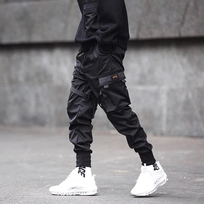 Schwarze Gothic Techwear Cargo-Hose getragen von männlichem Model gegen Betonwand, zeigt Seitenansicht mit Utility-Taschen-Details, matter Stoff-Textur und schwarzer Styling-Kombination mit weißen Sneakern