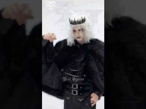 Männliches Model trägt kompletten schwarzen Gothic Umhang-Kostüm mit Krone auf dunklem Hintergrund, zeigt theatralischen Goth-Stil