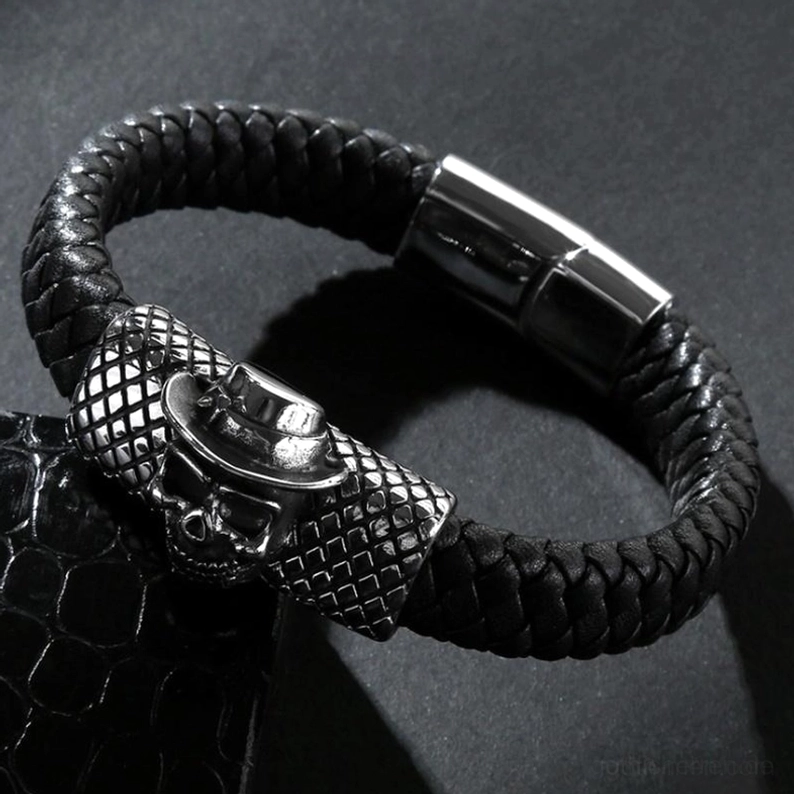 Dreiviertelperspektive des schwarzen gewebten Leder Schädel Armbands mit Zylinderhut-tragendem Schädel-Ornament und Magnetverschluss-Schnalle auf grauem Hintergrund