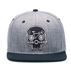 Frontalansicht der Skull Ride Hard Gothic Snapback Kappe, zeigt detailliert das Totenkopf-Fahrrad-Emblem und RIDE HARD Text auf Heather-Grau-Krone mit schwarzer flacher Krempe, 6-Panel Konstruktion und Belüftungslöcher