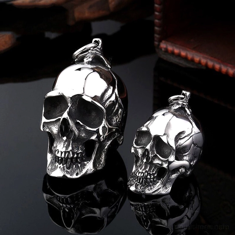 Paar dunkle Totenkopf-Anhänger aus Edelstahl mit detaillierter Knochenschnitzerei und Spiegelreflexion auf schwarzem Untergrund