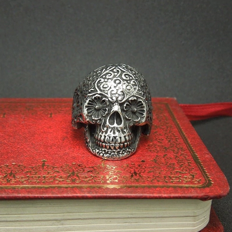 Ornamental gravierter Totenkopfring auf vintage rotem Buch präsentiert, antike dunkle silberne Edelstahl-Finisch, vollständige florale Engravierungsabdeckung, literarische Gothic-Asthetik-Styling