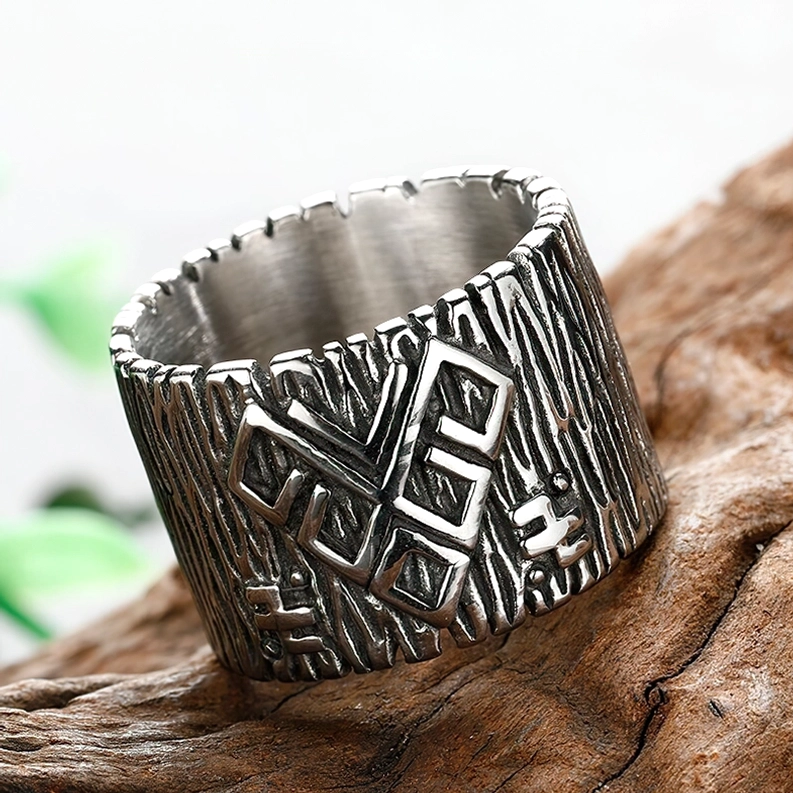 Dreiviertelansicht des breiten Band-Runen-Rings mit vintage Silberton und eingeprägten Runensymbolen vor Holzoberfläche mit grünem Laubhintergrund, gotische Ästhetik